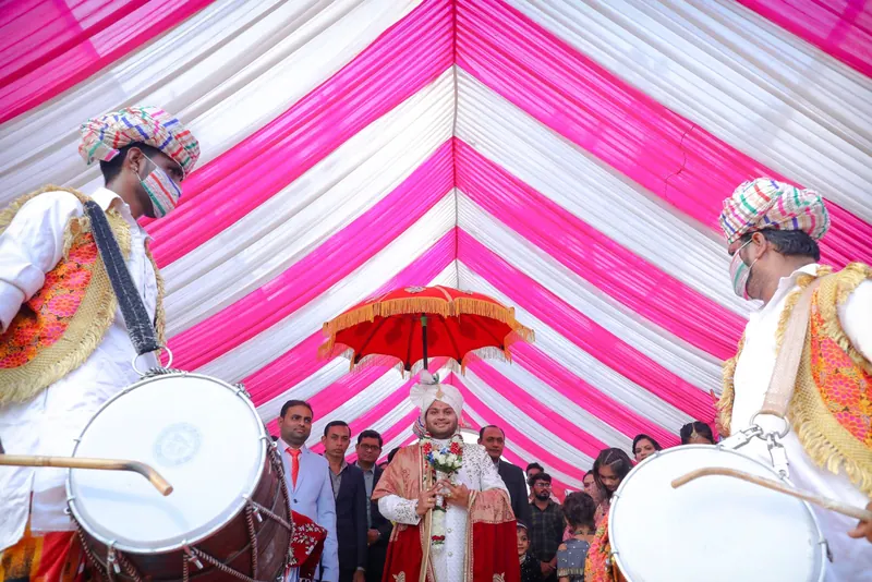 Dhol players drumming as the groom arrives under a red umbrella beneath a pink-and-white canopy