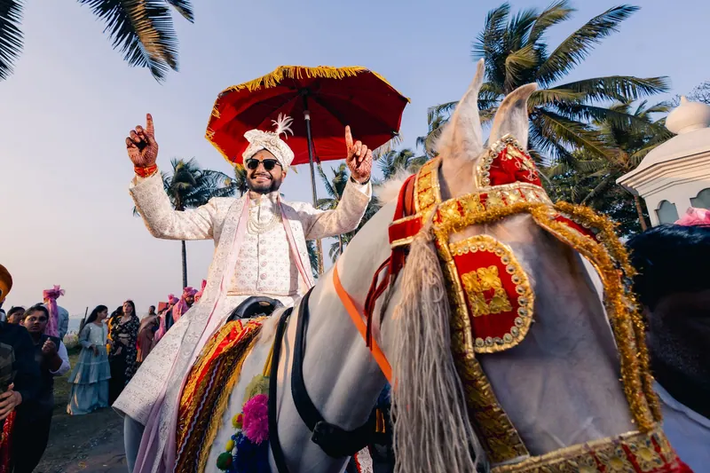 Joyful groom in an ivory sherwani riding a decorated horse beneath a red umbrella during the baraat