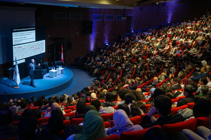 Tiered auditorium full of delegates watching a speaker on a blue-lit round stage