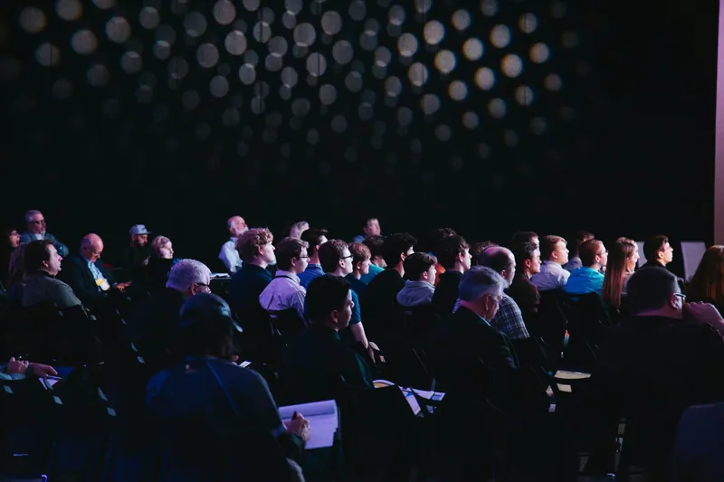 Conference audience seated in a dark hall beneath a dotted lighting ceiling
