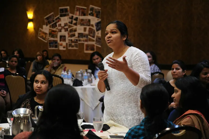 Indian woman in white speaking into a microphone at a seminar with women seated at tables