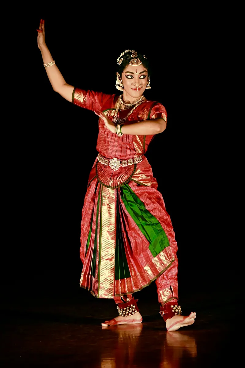 Bharatanatyam dancer in a red silk costume mid-pose under a spotlight