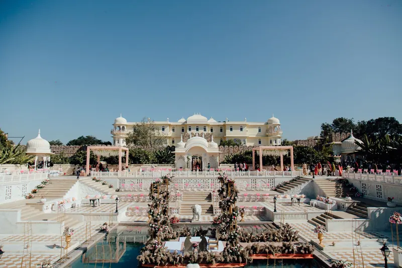Palace venue with a floral mandap on a lily pool, white pavilions and stepped terraces under a clear sky