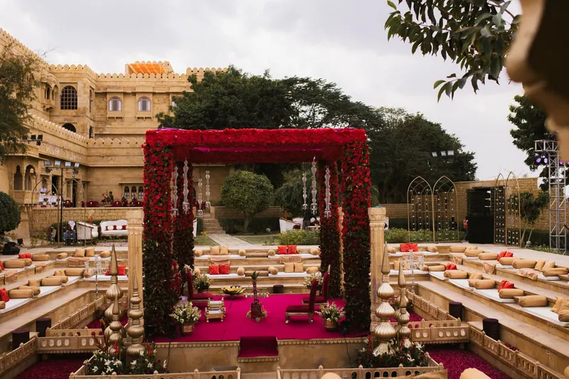 Red-rose mandap set in a sandstone palace courtyard with low floral seating for guests