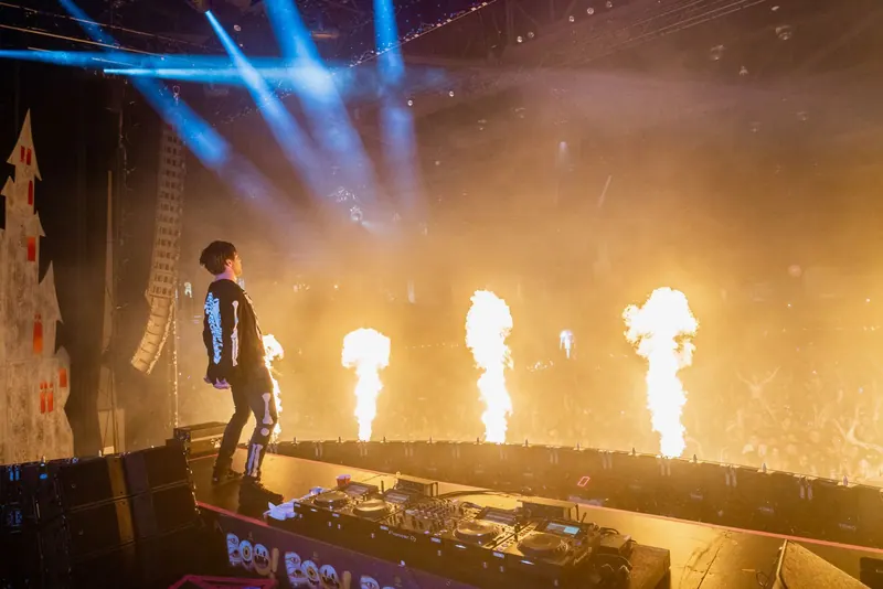 Performer on stage above DJ decks as flame jets fire across a hazy, lit arena