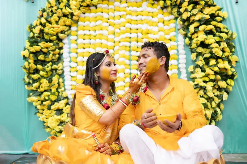 Bride applying haldi to the groom's cheek in front of a yellow-and-white marigold backdrop