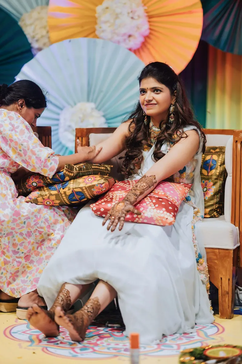 Mehendi artist applying henna to the bride's hands and feet beside colourful paper-fan decor