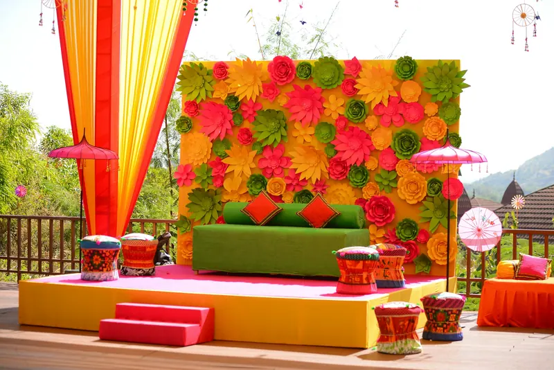 Colourful outdoor mehendi stage with a floral wall, green sofa and bright drapes set against the hills
