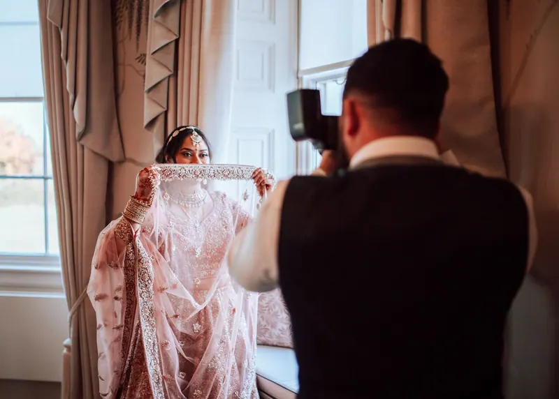 Photographer shooting a bride in a blush lehenga beside a window