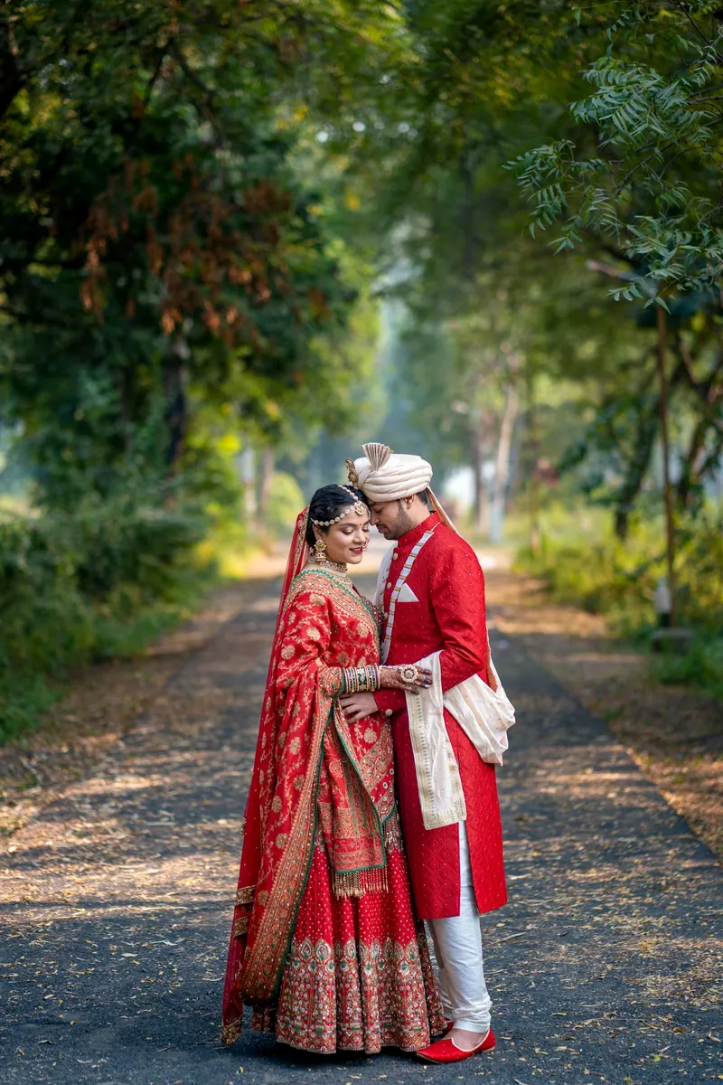 Couple in bridal red and a matching sherwani embracing on a tree-lined forest path