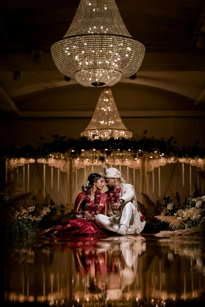 Newlyweds seated on a glowing stage beneath tiered crystal chandeliers, reflected in a polished floor