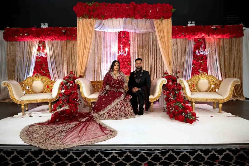 Couple posing on a red-and-gold reception stage with ornate gold chairs and floral canopies