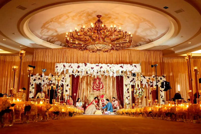 Wide view of a grand ballroom wedding with a floral mandap stage beneath a crystal chandelier and candles lining the aisle