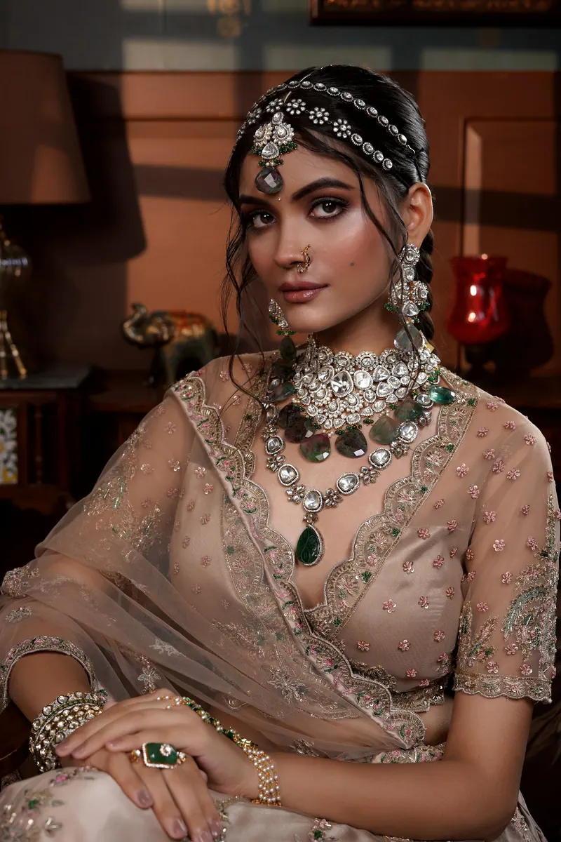 Bride in a peach embroidered lehenga and emerald-and-diamond necklace seated in a softly lit room