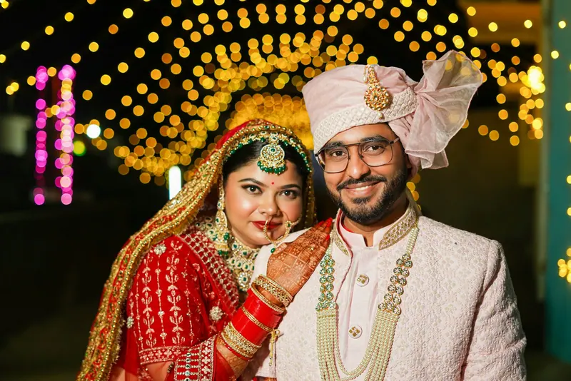 Bride in a red lehenga and groom in an ivory safa posing cheek to cheek beneath an arch of golden fairy lights