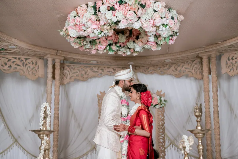 Bride in red and groom in an ivory sherwani sharing a quiet moment under a carved white mandap with a hanging floral chandelier