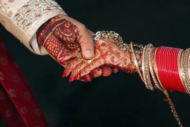Groom holding the bride's hennaed hand, adorned with red chooda bangles and gold kaleere, against a dark background