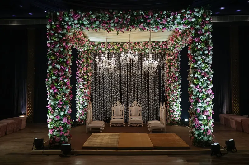 Floral mandap wrapped in pink and white blooms with crystal chandeliers and ornate white chairs inside a darkened hall