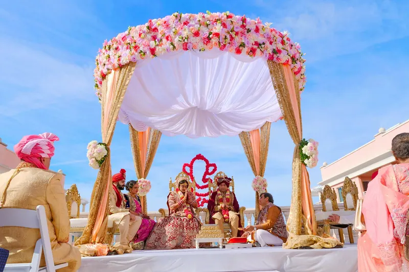 Couple seated beneath a pink-and-white floral canopy mandap at an outdoor ceremony under a bright blue sky