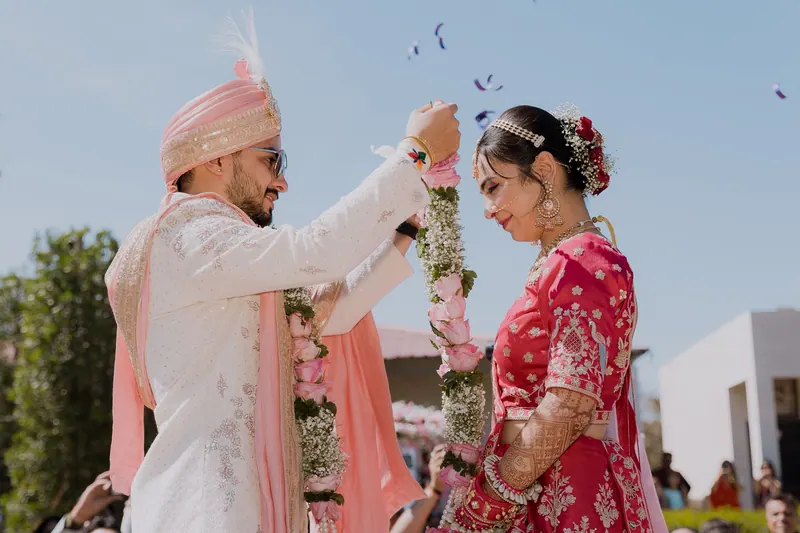 Groom in a pink safa placing a flower garland on the bride during an outdoor varmala under a blue sky