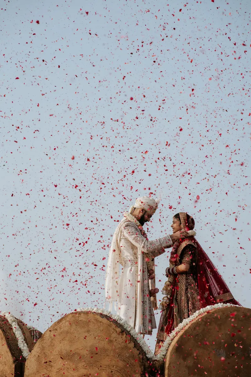 Bride and groom exchanging garlands, raised high as rose petals rain down against a pale sky